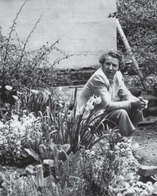 Capi as an older woman sitting in her garden, turned away from the camera, looking over her shoulder as if speaking to someone off-camera