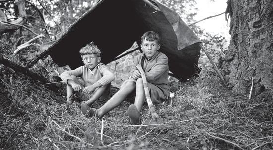Two young boys sitting beneath a shelter made of sticks and a tarp