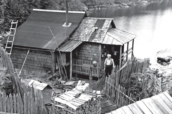 A wooden house with a man in a cap and suspenders standing before it