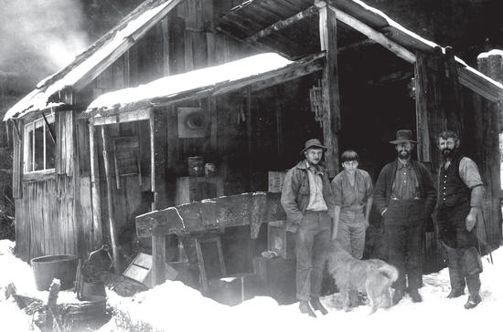 A group of four (one woman, three men) and a dog standing in the snow outside a wooden building