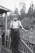 An older man frowning and standing with his hand on a fencepost in front of a house