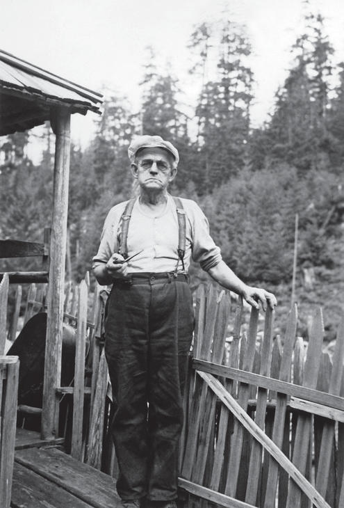 An older man frowning and standing with his hand on a fencepost in front of a house