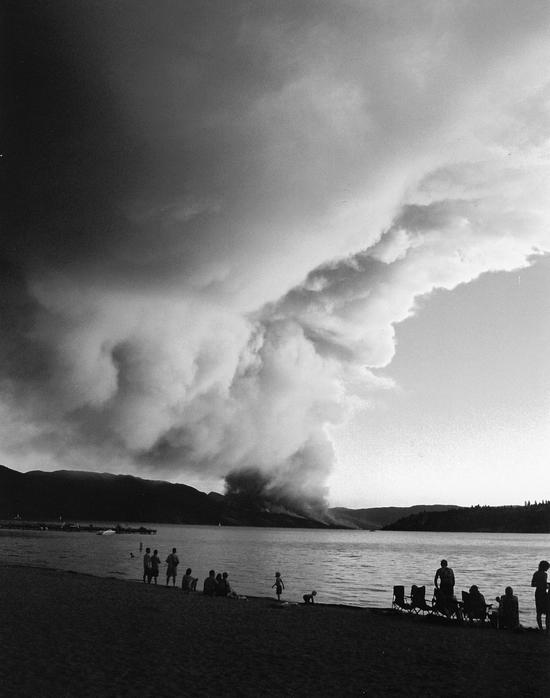 A group of people on a beach watch a large plume of smoke in the distance.