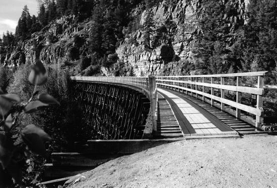 A bridge with railings supported by wooden beams. The bridge is built by a cliffside and connects two pieces of land separated by a dip. 