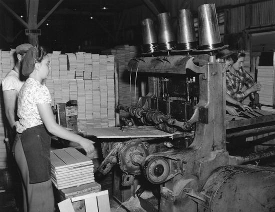 Two people standing in front of a machine indoors. One person feeds wood planks into the machine. Behind them are tall piles of wood planks.
