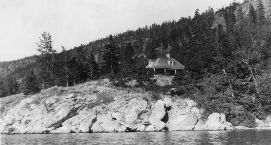 A house surrounded by trees built on a short cliff leading to a body of water. The photo is shot from the water.