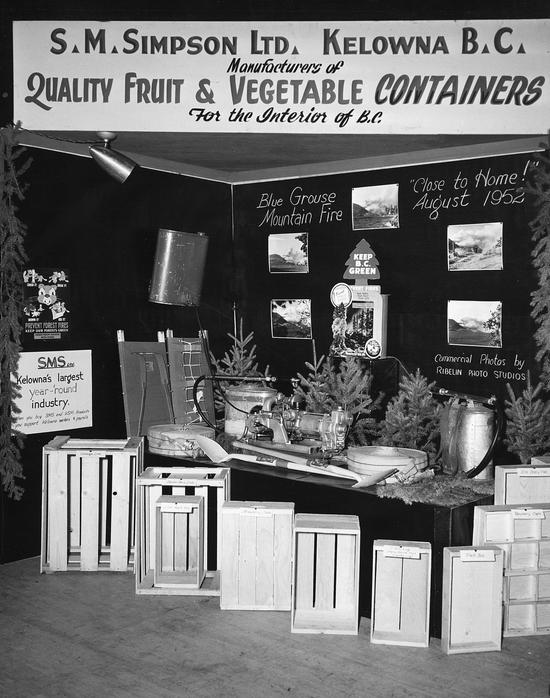 A display including a table holding multiple devices and tree clippings. In front of the table are labelled wooden boxes and behind the table are images of scenery. 