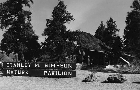 A wooden sign in front of an outdoor roofed area surrounded by trees. Text: Stanley M. Simpson Nature Pavilion