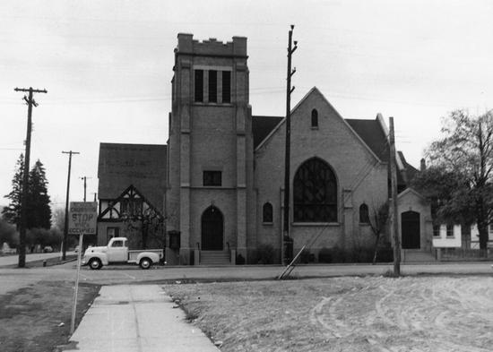 The view of a large church with large gothic windows from across the street. A pickup truck is parked at the church.