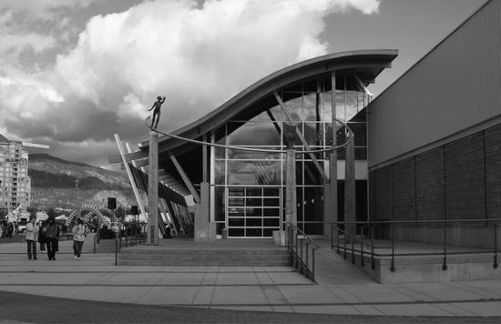 A long building with a curved roof and walls made of windows. Before it is a statue of a head and one of a person reaching out. To the left is a large paved walkway where people are walking.