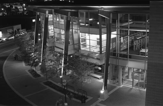 A picture at night of a large building with walls made of windows. Shelves of books can be seen within. Outside is a path made of brick and small trees. 