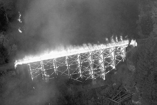 A bridge on fire. The bridge is supported by wooden beams and is connecting two pieces of land separated by a dip. 