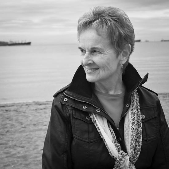 A portrait of a smiling woman with short hair wearing a leather jacket and a scarf. Her body is facing the camera but her head is turned to look to the left. She is standing on a beach.