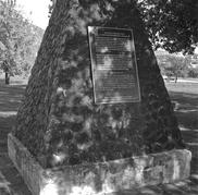 A structure made of stone in the vague shape of of pyramid. On it is a plaque with indiscernible text. In the background are trees.