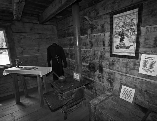 The inside of a wooden building. There are several items in frame including a small table holding a book, a chest, cooking pans, a picture and plaques. In the corner stand dark robes. On the robes, where a person's neck would be, hangs a large cross.