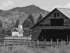 A wooden building stands in a field, some of its planks fallen off. Behind the building is a light-coloured church with a cross on top of its roof. In the background are hills and trees.