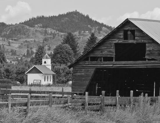 A wooden building stands in a field, some of its planks fallen off. Behind the building is a light-coloured church with a cross on top of its roof. In the background are hills and trees.
