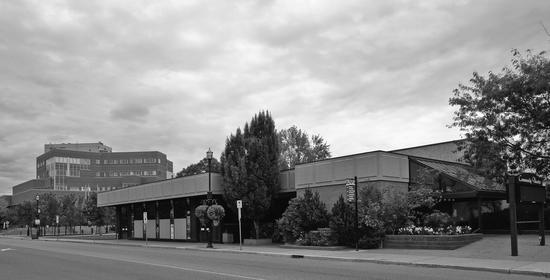 A short building within a city. The building is surrounded by flower beds, bushes and trees.