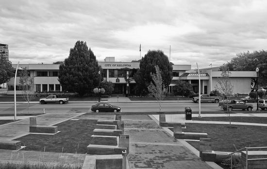 Doors to a large building with two storeys framed by trees as viewed from across a road. Modern cars are parked on the sides of the road. Text: City of Kelowna