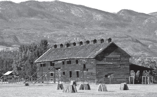 A large old wooden building stands in a field behind hay bales and in front of trees and hills.