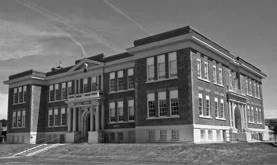 A large school covered in windows stands surrounded by grass, trees and bushes. Its entry ways have arches and pillars.