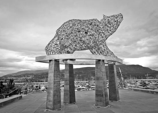 A statue of a bear suspended above the ground. The bear is made of sticks connected to form geographical shapes. It has various items scattered within it including fruits and smaller bears.