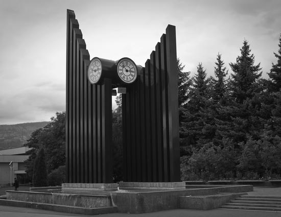 Two clock faces pointed in different directions. They are held into the air by two groups of ten poles. The poles are lined up in descending height order and form two walls.
