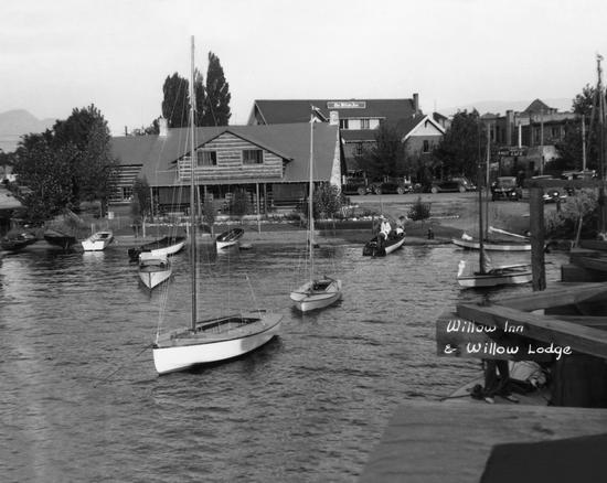 Boats floating on a body of water a viewed from a dock. Behind them is a wooden building.