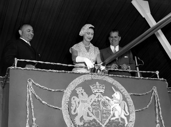 A smiling woman wearing a dress, a hat and gloves is about to cut a large ribbon. Two men in suits stand on either side of her, watching with smiles. They all stand on a platform decorated with a crest.