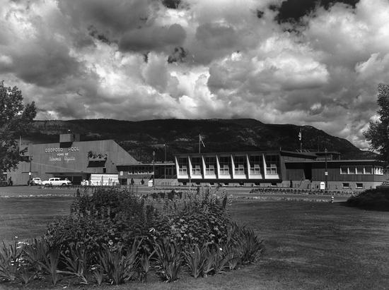 A long building. In front of it is a large area with a driveway for cars. It also houses grass, flowers, trees and shrubbery. Text: Ogopogo Pool Home of Kelowna's Regatta