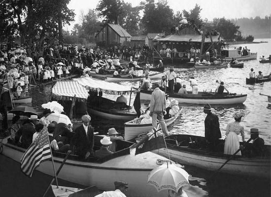 Numerous boats. Some boats are docked while some are pulled away from the dock. A crowd of people watch from the dock, a nearby building and stands for viewing. 