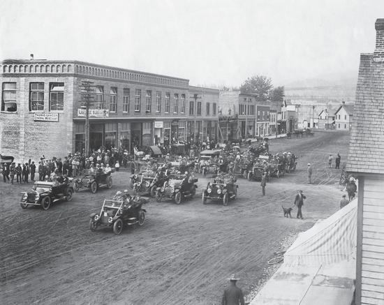 An older dirt road surrounded by shopfronts. A small crowd watches as a group of old cars drive by.