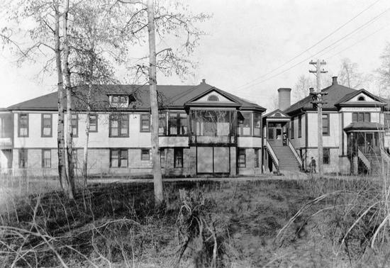 A large, light-coloured building with dark trimming. A staircase leads to an entrance. 