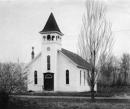 A church framed by trees. A bell and a cross sits on its roof.