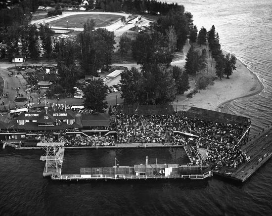 An aerial view of a sectioned off portion of a body of water. Behind it are stands filled with people and a large building. Further past this are trees and pathways leading to a field.