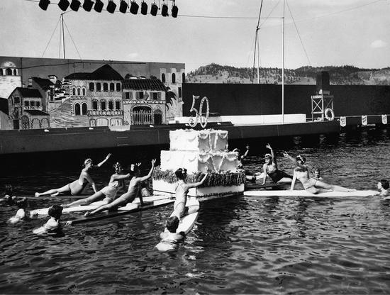 A large two-tiered cake displaying the number 50 floats on a body of water. Around it, women reach toward it whilst on boards held by another set of women in the water. 