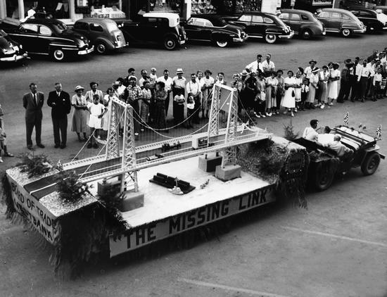 An old car pulls a platform with a model of a bridge. A crowd of people watch to the side, and behind them a line of old cars are parked.