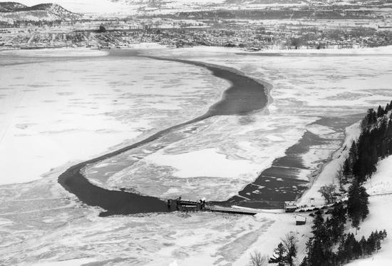 A lake covered in ice. There is a path connecting two pieces of land that is uncovered by ice. There is a dock at each end of the path.