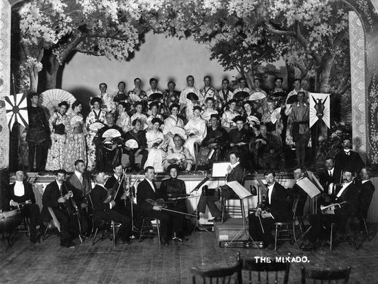 A large group of people sit and stand on a stage wearing Japanese-styled clothing while holding fans. Surrounding them are fake trees and in front of them, below the stage, is an orchestra. Text: The Mikado