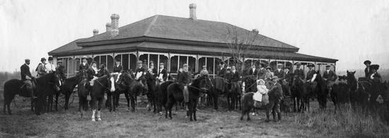 A group of people sitting on horses in front of a one-story building covered in windows.