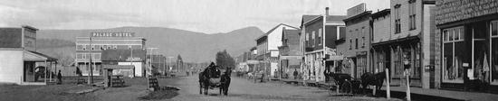A street lined with shops. A person in a horse-drawn cart rides down the street while another horse-drawn cart is parked at a shop.