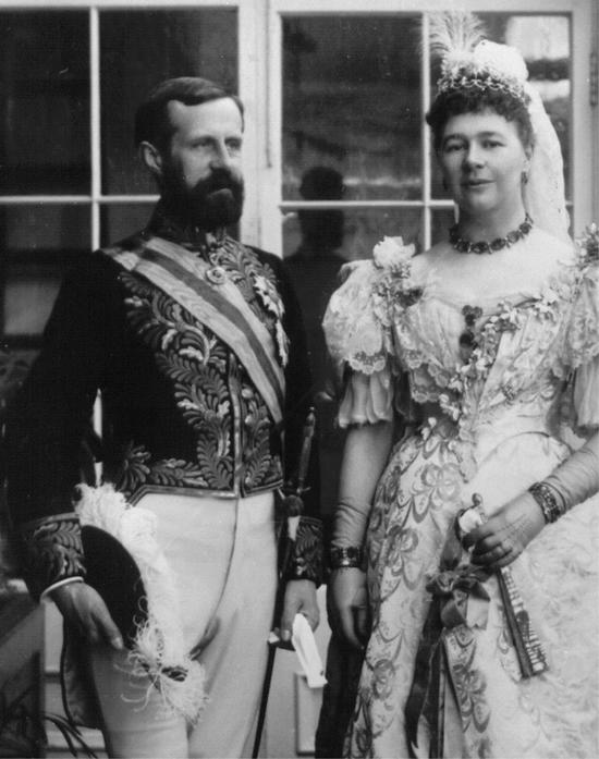 A man and woman stand posed in old formal wear. The woman looks into the camera while the man looks facing to the right. They are in front of windows.