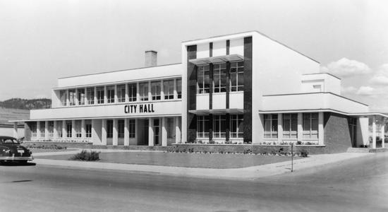 The front of a large light-coloured building with two storeys. An old car is parked on the road in front of the building. Text: City Hall
