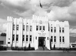 A large light-coloured two storey building with a flag on its roof and a clock above its entrance. Text: Post Office Kelowna