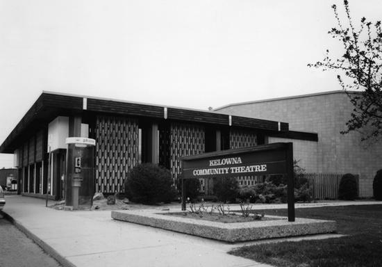 A small building made of wood with a sign standing by its side. Text: Kelowna Community Theatre