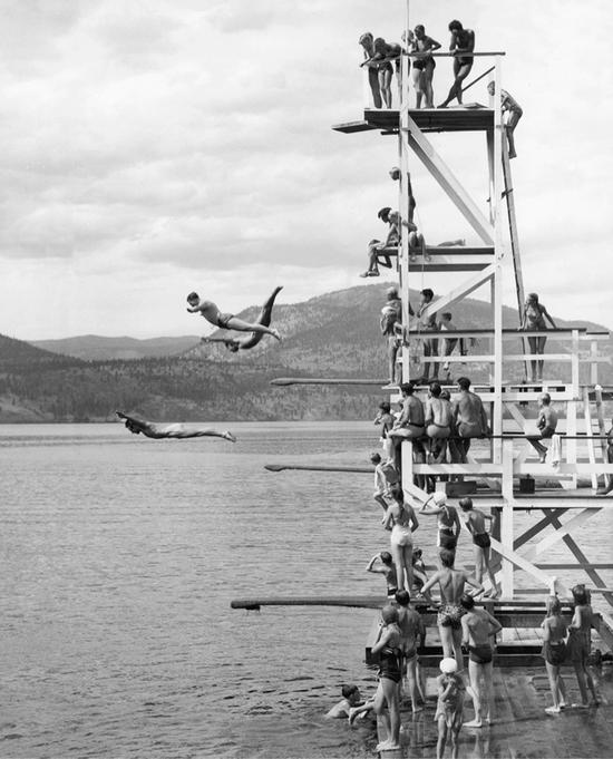 A wooden diving tower with five levels carries a large group of people. They watch as three people fall toward the water below.