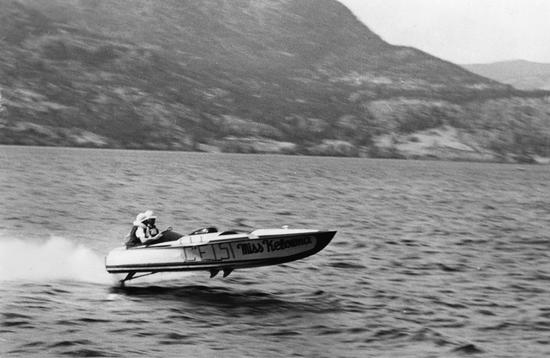 A speedboat racing over a body of water, its speed lifting it into the air as water sprays in its wake. The hills in the background are blurred by the rapid motion.