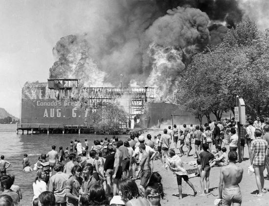 A large structure built atop water is on fire, billowing smoke. Part of its framework is exposed. A crowd watches from a nearby beach, many people in swimsuits.