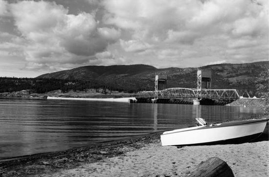 A bridge as seen from a nearby beach with a small boat lying on the sand. In the background are tree-covered hills.