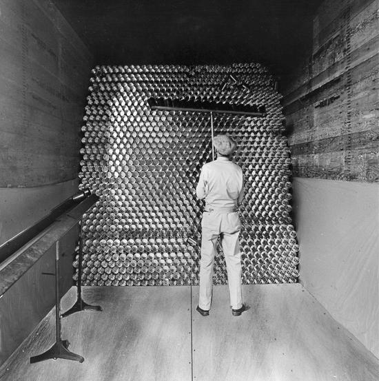 A man stands in a narrow space facing a tall stack of cans. He holds a tool to help maneuver the cans into place.